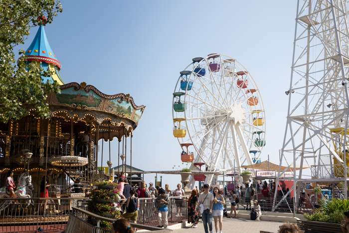 The scene captures a lively amusement park bustling with people enjoying various attractions. On the left, a beautifully ornate carousel with a colorful canopy features intricately painted scenes and bright lights, while several riders can be seen aboard the carousel. To the right, a large Ferris wheel towers above the crowd, adorned with vibrant passenger cabins in multiple colors. Groups of visitors are scattered throughout the area, some walking close to the carousel, while others line up near food stalls marked by brightly colored awnings. The clear blue sky and lively atmosphere suggest a perfect day for enjoyment and entertainment.