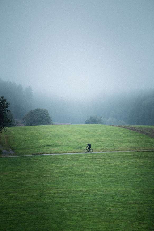 A lone cyclist rides along a narrow path through an expansive green field. The surroundings are enveloped in a thick mist, creating a serene and slightly mysterious atmosphere. In the background, the contours of trees and distant hills are barely visible through the fog, enhancing the sense of solitude and tranquility.