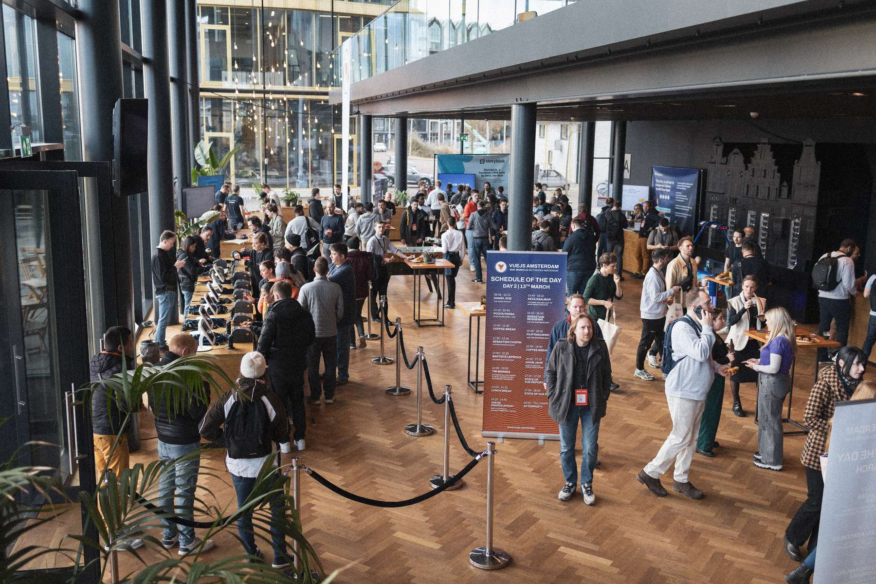 A bustling scene inside a modern venue filled with attendees engaging in discussions and networking. Groups of people gather around tables, some enjoying refreshments while others stand in conversation. Large windows allow natural light to illuminate the space, showcasing natural wood flooring and contemporary design elements. A sign displaying the day's schedule is prominently featured, and various digital displays can be seen in the background. Lush green plants add a touch of nature to the vibrant atmosphere.