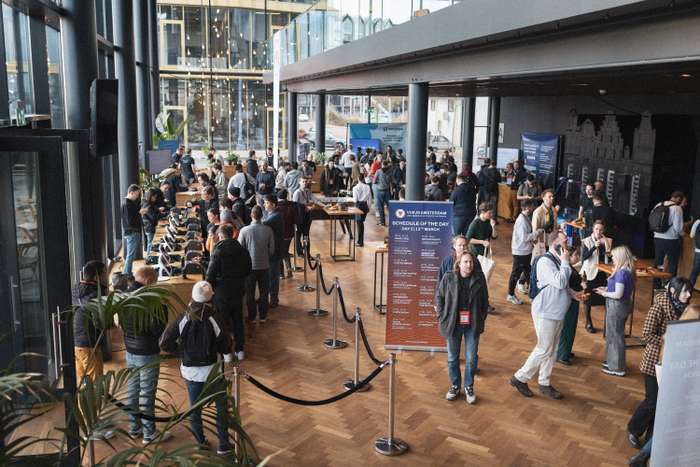A bustling scene inside a modern venue filled with attendees engaging in discussions and networking. Groups of people gather around tables, some enjoying refreshments while others stand in conversation. Large windows allow natural light to illuminate the space, showcasing natural wood flooring and contemporary design elements. A sign displaying the day's schedule is prominently featured, and various digital displays can be seen in the background. Lush green plants add a touch of nature to the vibrant atmosphere.