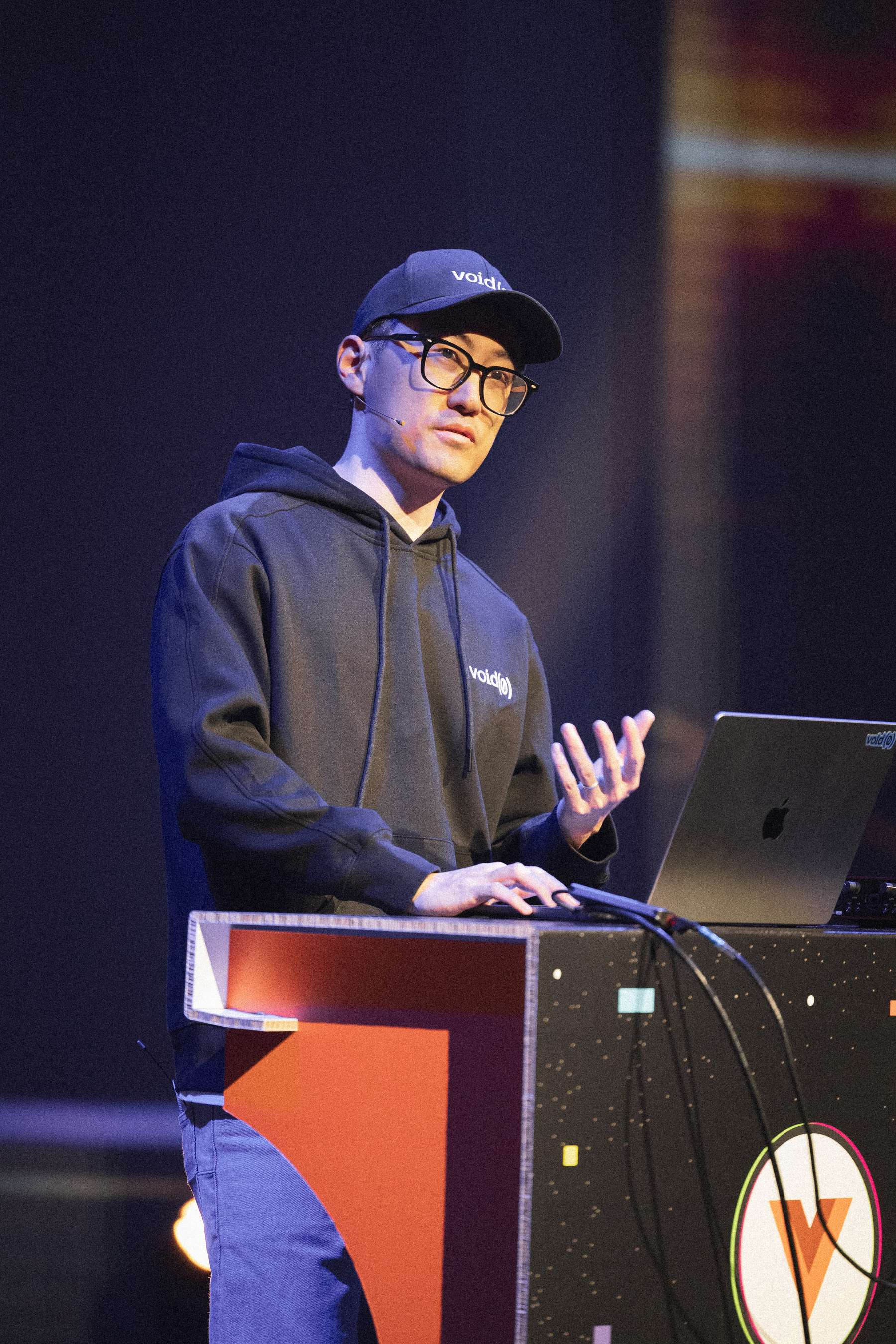 The speaker Evan You stands at a podium with a sleek laptop and audio equipment, wearing a black hoodie and a cap. The background features a colorful, blurred gradient, adding a dynamic atmosphere to the scene. The speaker appears focused and animated as he gestures, engaging with the audience, conveying a sense of enthusiasm about the topic being discussed.