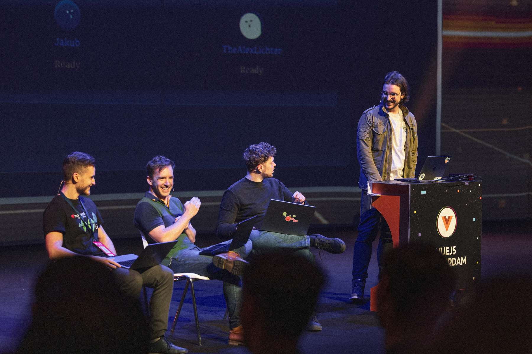 A lively scene from Vue.js Amsterdam featuring four speakers on stage (Jakub Andrzejewski, Daniel Roe, Alexander Lichter and Alvaro Saburido Rodriguez). The first three speakers are seated on white chairs, dressed in casual attire, with one individual smiling while looking at the audience. The fourth speaker stands behind a podium with a colorful design, energetically engaging the audience. Each speaker has a laptop in front of them, indicating a presentation or demonstration. A large screen in the background displays usernames and a status message, contributing to the interactive environment.