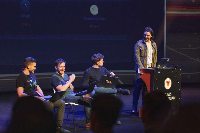 A lively scene from Vue.js Amsterdam featuring four speakers on stage (Jakub Andrzejewski, Daniel Roe, Alexander Lichter and Alvaro Saburido Rodriguez). The first three speakers are seated on white chairs, dressed in casual attire, with one individual smiling while looking at the audience. The fourth speaker stands behind a podium with a colorful design, energetically engaging the audience. Each speaker has a laptop in front of them, indicating a presentation or demonstration. A large screen in the background displays usernames and a status message, contributing to the interactive environment.