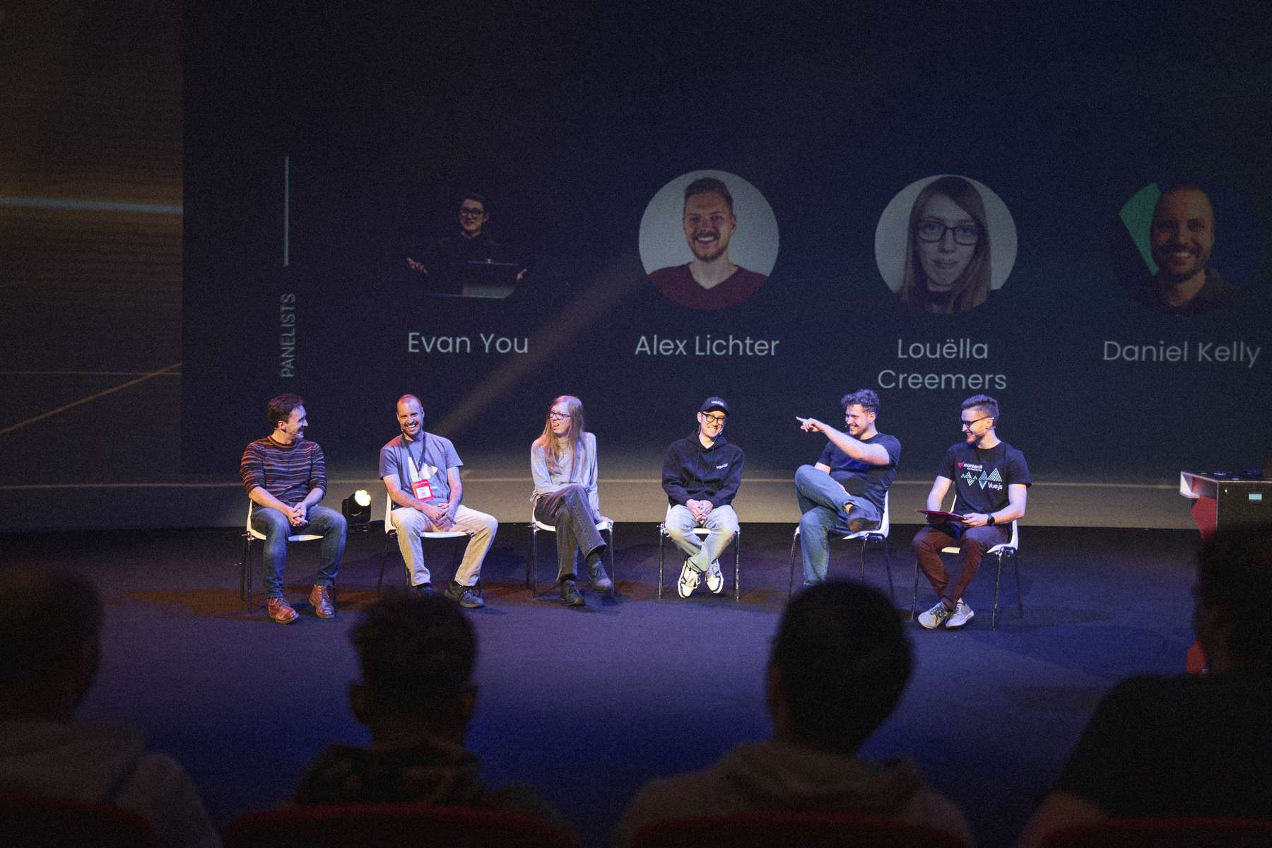 A panel of six individuals (Daniel Roe, Daniel Kelly, Louëlla Creemers, Evan You, Alexander Lichter and Jakub Andrzejewski) is seated on stage at Vue.js Amsterdam, engaging in discussion during the panel discussion. Each panelist appears to be sharing insights, with two of them actively gesturing. The background features names and images of the panelists displayed prominently, enhancing the setting. The audience is partially visible, seated in the foreground, indicating an interactive environment. The lighting is focused on the panel, creating an inviting atmosphere for conversation.