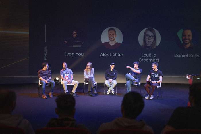 A panel of six individuals (Daniel Roe, Daniel Kelly, Louëlla Creemers, Evan You, Alexander Lichter and Jakub Andrzejewski) is seated on stage at Vue.js Amsterdam, engaging in discussion during the panel discussion. Each panelist appears to be sharing insights, with two of them actively gesturing. The background features names and images of the panelists displayed prominently, enhancing the setting. The audience is partially visible, seated in the foreground, indicating an interactive environment. The lighting is focused on the panel, creating an inviting atmosphere for conversation.