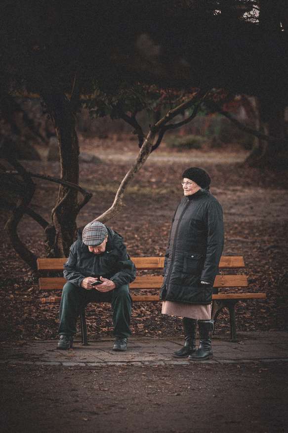 An elderly couple experiences a quiet moment in a park with the man seated on a wooden bench looking at his mobile phone, while the woman stands beside him looking away thoughtfully. A tangle of bare, dark branches creates a moody backdrop, suggesting the late autumn or winter season. The scene conveys a sense of stillness and the complexity of human relationships.