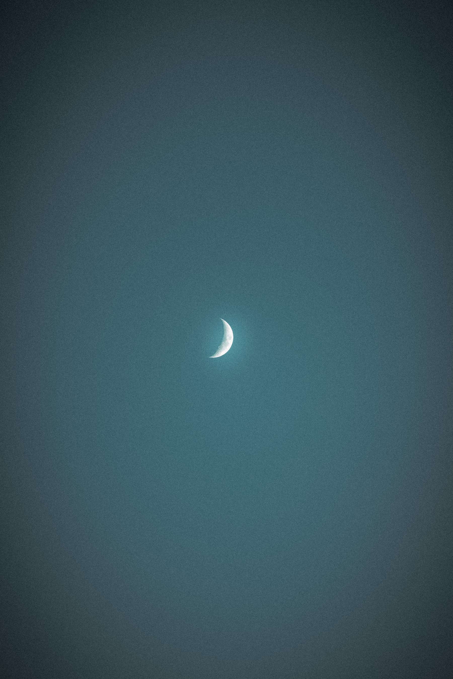 The image captures a waxing crescent moon centered in a dark, textured night sky. The moon is brightly lit on its curved edge, fading into darkness toward the rest of its surface, which is barely visible against the backdrop of the evening glow.