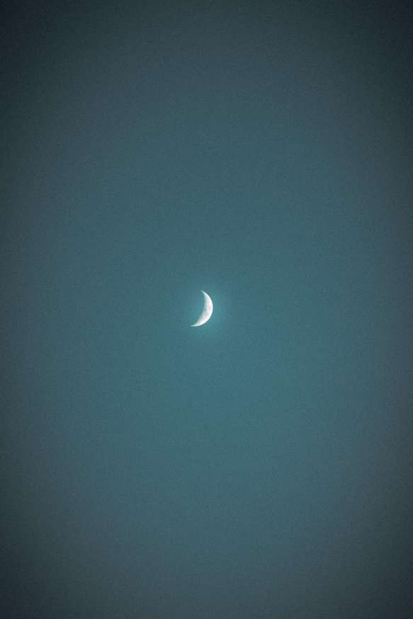 The image captures a waxing crescent moon centered in a dark, textured night sky. The moon is brightly lit on its curved edge, fading into darkness toward the rest of its surface, which is barely visible against the backdrop of the evening glow.