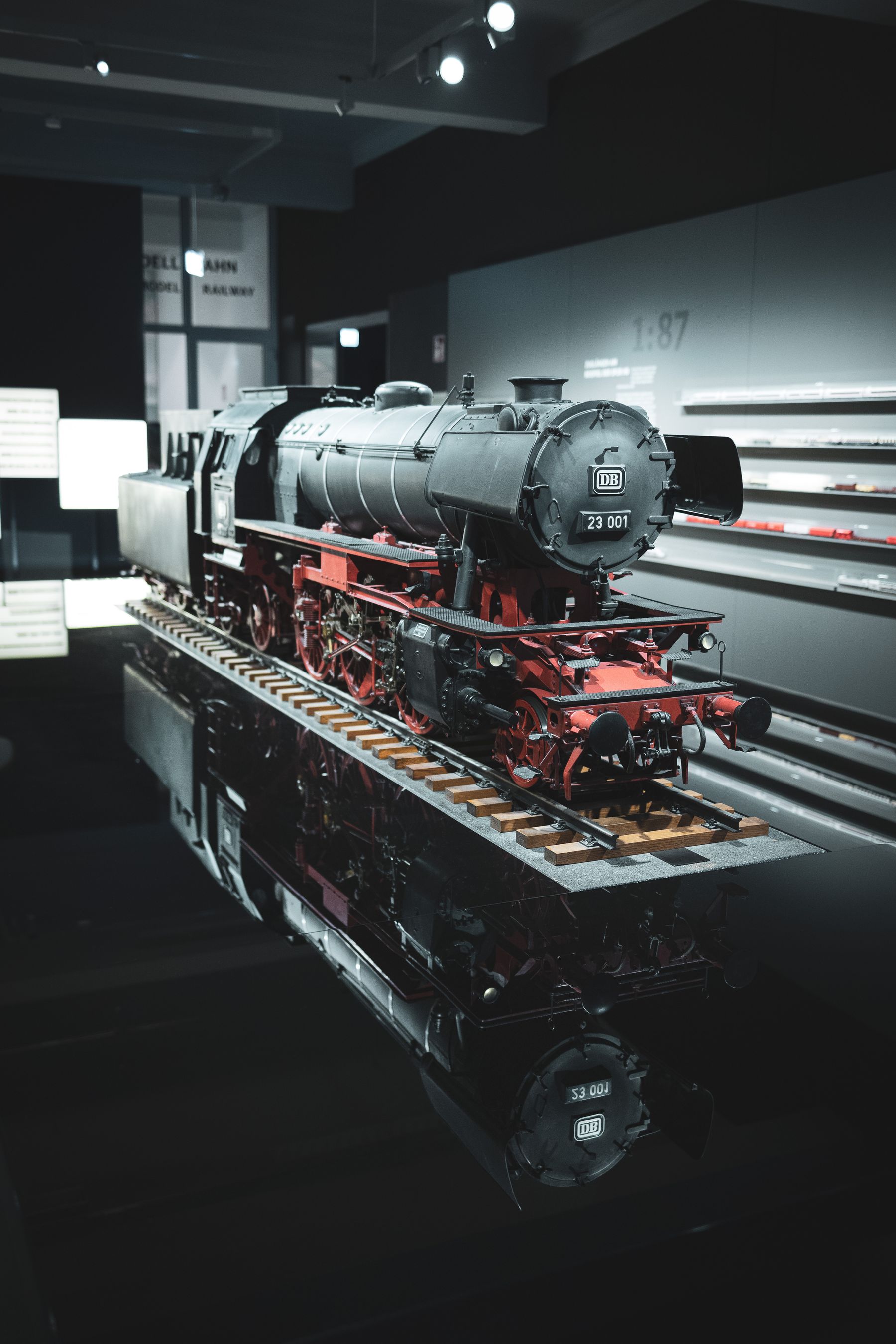 The image showcases a detailed scale model of a classic steam locomotive, predominantly black with red trimmings, displayed on tracks. The model is identified as '23 001' marked on the locomotive's front, with the 'DB' logo on the front. The setting appears to be an exhibition with carefully directed lighting highlighting the train model and creating a reflective surface below it. In the background there are informational displays and subdued lighting, suggesting an indoor museum environment.
