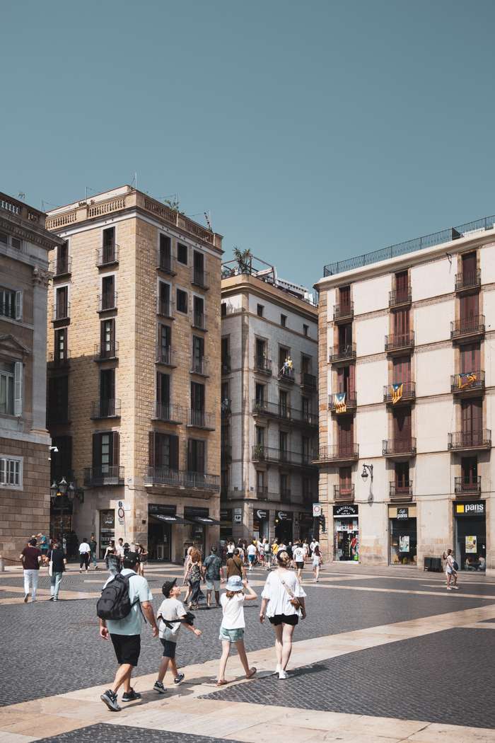 A vibrant city square filled with people enjoying a sunny day. Families and tourists walk along the cobblestone pavement, admiring the charming architecture of the surrounding buildings. The structures exhibit a mix of styles, with balconies adorned with colorful flags and greenery on rooftops, set against a clear, blue sky. Shops line the street, enhancing the lively atmosphere as pedestrians engage in leisurely activities.