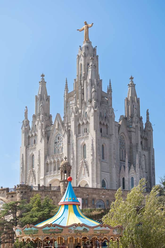 In a serene landscape, a grand cathedral with intricate Gothic architecture towers prominently, crowned by a golden statue with outstretched arms. This impressive building features tall spires, ornate stonework, and a large rose window, reaching towards a clear blue sky. In the foreground, a vibrant carousel with a colorful striped canopy, adorned with fairy lights and classic artwork, adds a playful contrast to the historical monument, inviting visitors to enjoy the moment.