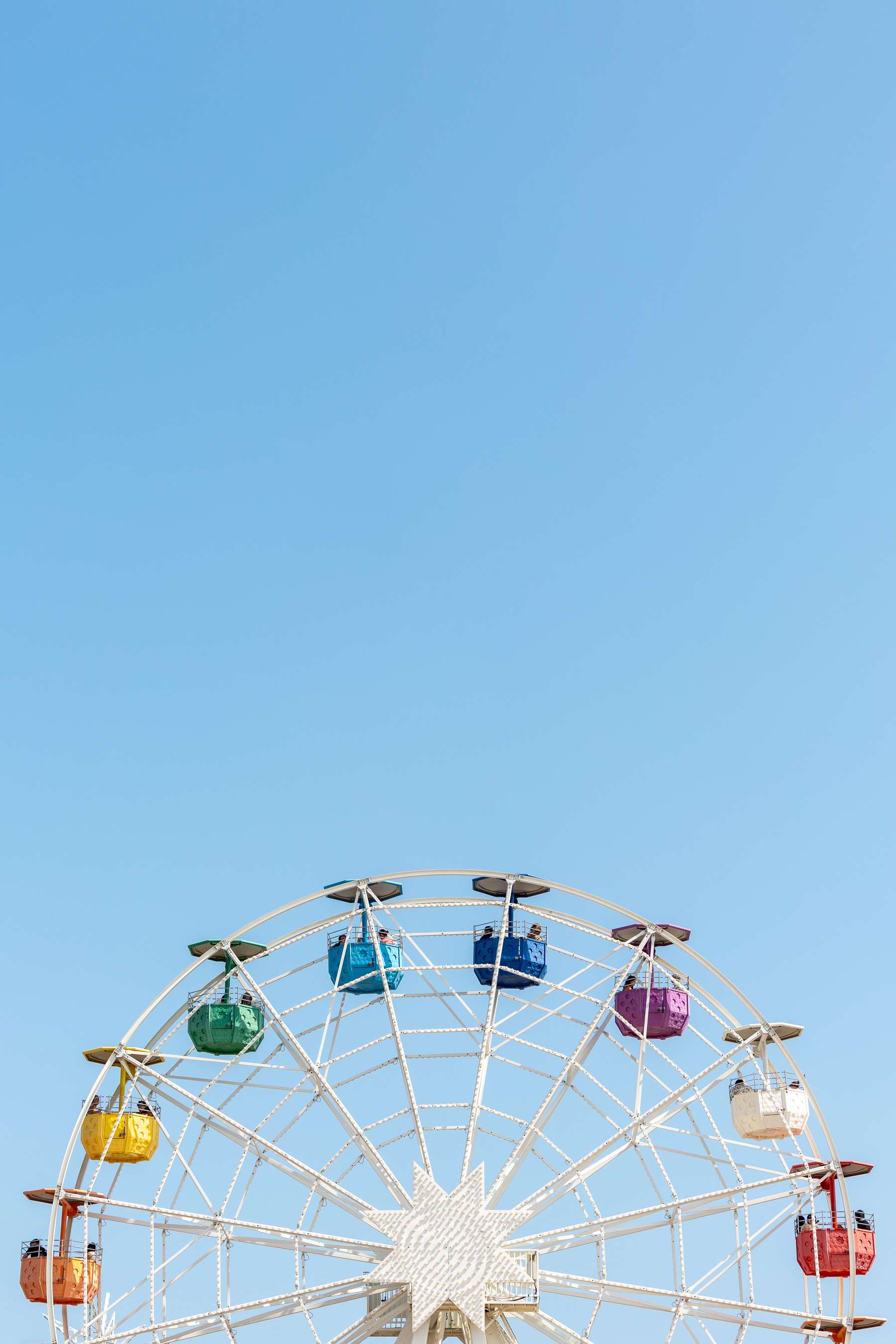 A colorful Ferris wheel stands against a clear blue sky, featuring bright cabins in various shades including yellow, green, blue, purple, red, and white. The structure is intricately designed with a white frame, and a large star shape at the base, creating an inviting atmosphere for visitors.