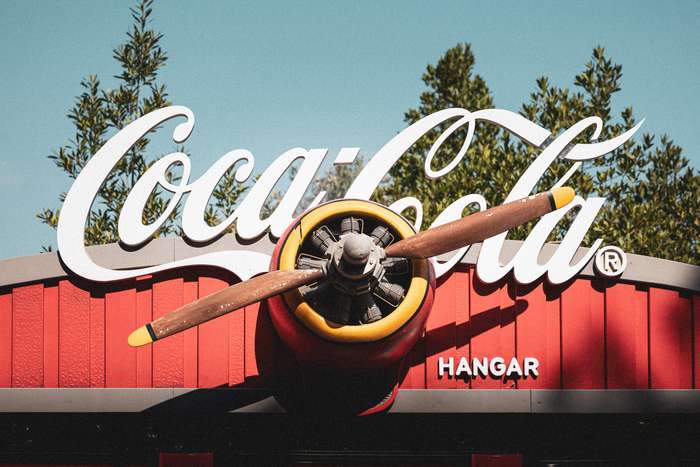 The scene features a striking display of a Coca-Cola sign with a vintage aviation theme. At the center, a large, round propeller, painted in yellow and brown, protrudes from a red backdrop. The bold Coca-Cola logo, in white cursive lettering, arches above the propeller, while the word 'HANGAR' is prominently displayed beneath it. The clear blue sky above contrasts with the vibrant colors of the sign and the lush greenery in the background, enhancing the overall visual appeal.