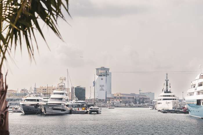 A bustling marina filled with several luxury yachts docked at the water's edge. In the background, a modern building stands tall, prominently displaying an advertisement for Louis Vuitton, while palm leaves frame the scene, adding a tropical touch. The sky is partially cloudy, casting a soft light over the water, creating a tranquil yet vibrant atmosphere. An airplane can faintly be seen in the sky, hinting at travel and movement in the busy harbor.