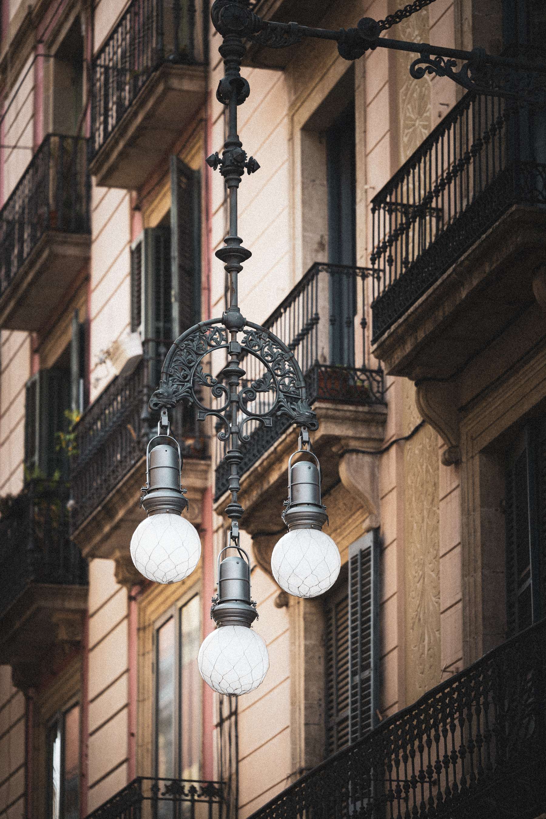 A beautifully designed street lamp hangs gracefully from an ornate iron fixture, featuring three decorative glass light bulbs. The background consists of a series of classic European-style balconies, with wrought iron railings and green shutters, set against a softly colored facade. The scene captures a blend of architectural elegance and warm ambient lighting, indicative of urban charm.