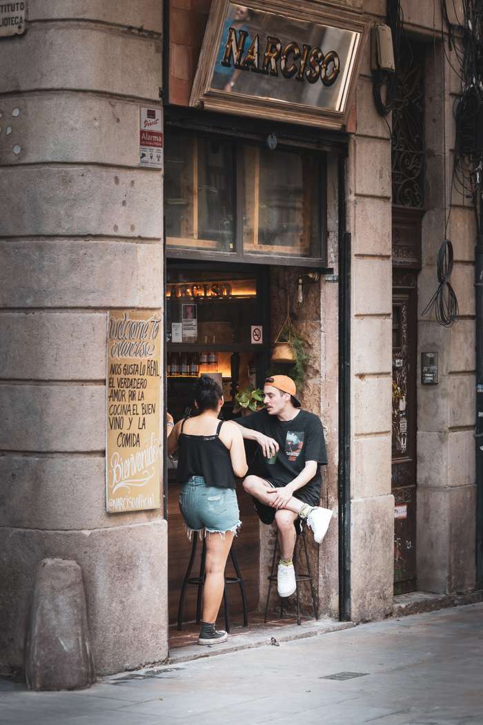 A young man and woman engaged in a conversation outside a café named 'Narciso'. The woman, wearing a black tank top and denim shorts, is seated on a stool, while the man, dressed in a black t-shirt and orange cap, is perched casually on another stool. The entrance of the café is framed by stone walls, with a welcoming sign in Spanish that invites patrons in. A warm ambiance is suggested by the dim lighting visible through large windows behind them, with potted plants adding a touch of greenery to the scene.