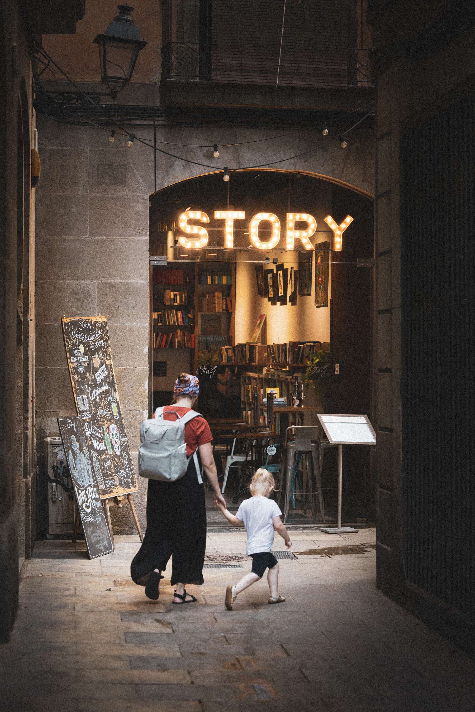 A woman, dressed in a red top and black skirt with a backpack, walks hand-in-hand with a young child down a cobblestone alleyway. The entrance to a cozy bookstore is visible ahead, with bright, marquee-style letters spelling "STORY" illuminated above it. A chalkboard menu stands to the side, decorated artistically, while the interior of the bookstore is glimpsed, filled with colorful books and a welcoming ambiance. The scene captures a warm, inviting moment between the two as they approach the shop.