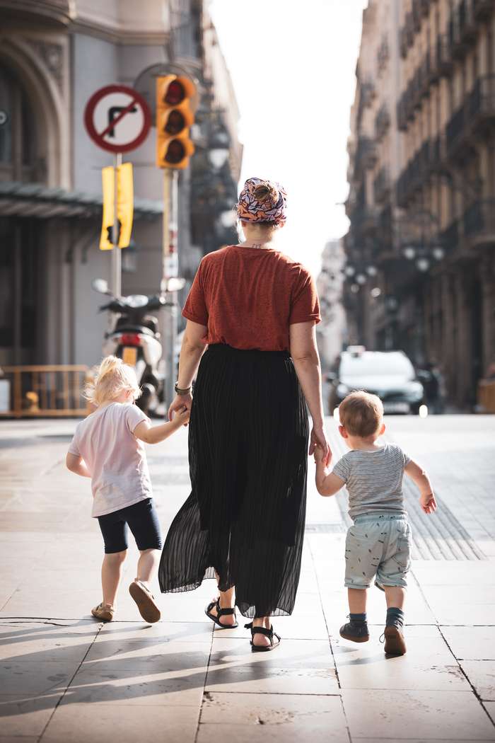 A woman walks hand in hand with two young children in an urban street, sunlight streaming down on them. The woman wears a rust-colored top and a black, flowy skirt, while her hair is styled with a colorful scarf. The older child, a girl with blonde hair, wears a light pink t-shirt and dark shorts, holding the woman’s hand. The younger child, a boy, is dressed in a gray striped shirt and light pants. In the background, there are street signs, a pedestrian crossing signal, and a blurred view of a car and buildings lining the street.