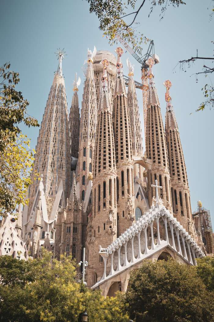 A close-up view of the Sagrada Família, showcasing its intricate towers reaching towards a clear blue sky. The detailed stonework features a variety of Gothic and Art Nouveau elements, with pointed spires adorned with colorful sculptures. Lush green foliage partially frames the scene, adding a natural touch to the architectural wonder, while construction detailing is visible at the top.