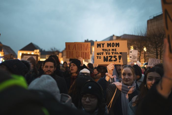 A diverse group of individuals are gathered in a twilight setting, participating in a protest. The crowd is dense and filled with various expressions of determination and focus. Several homemade signs are held aloft, with one prominently displayed in the center reading 'MY MOM TOLD ME NOT TO TALK TO NAZIS!' Other signs with messages are partially visible around it. The lighting suggests either dawn or dusk, and the urban setting provides a backdrop of buildings with the sky showing a gentle gradient from blue to orange.