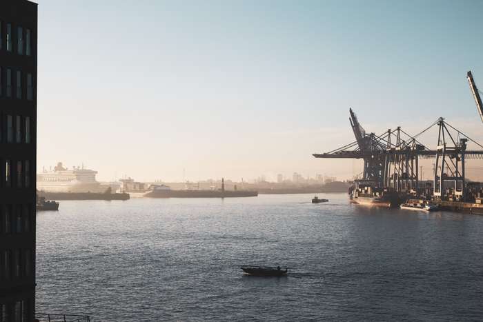 A tranquil harbor scene with calm waters reflecting soft morning light. In the foreground, a small boat glides across the water, while larger ships are docked along the shore. Cargo cranes loom in the distance, silhouetted against a hazy skyline, creating an industrial backdrop.