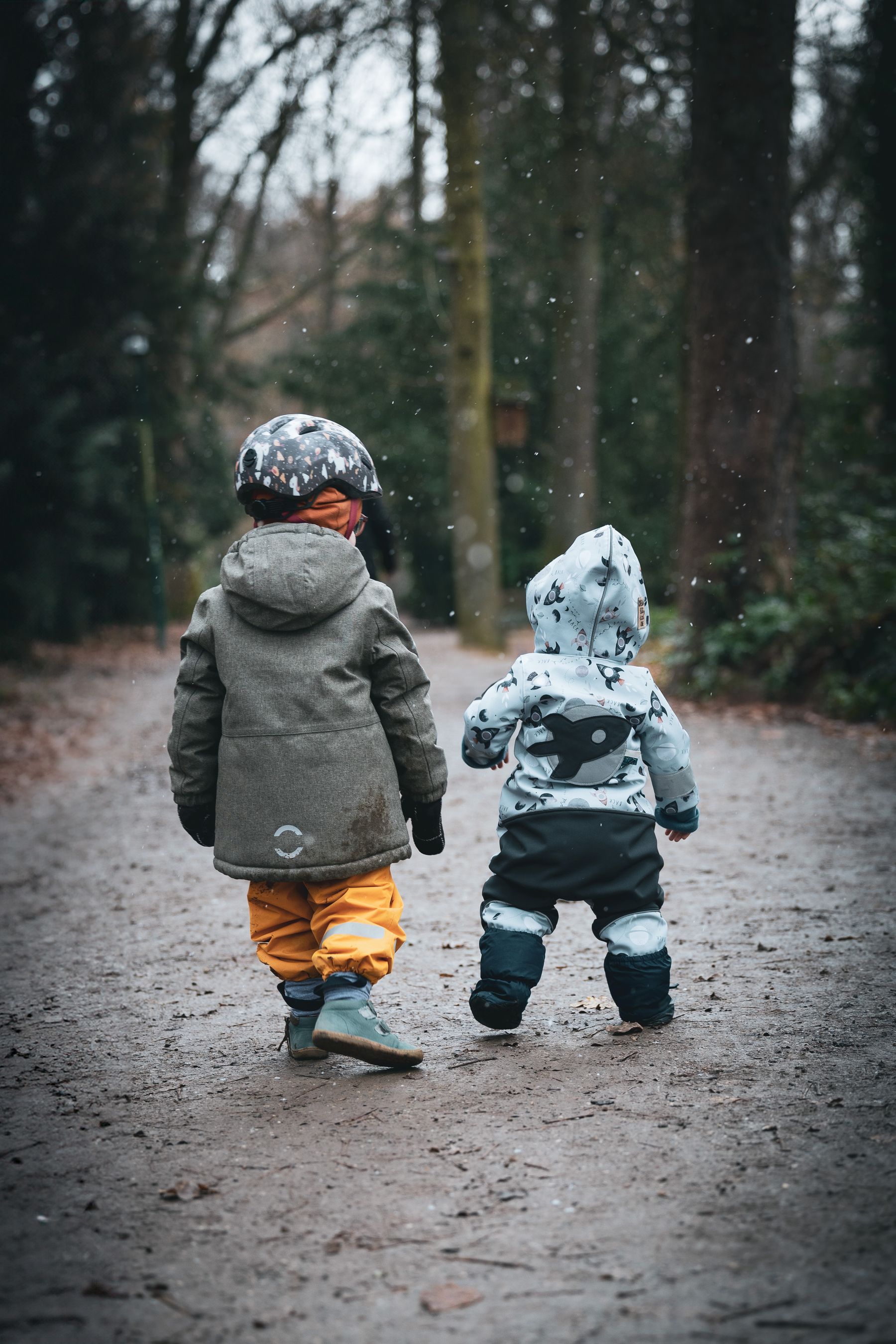 Two small children are walking away from the camera, hand in hand, on a dirt path amid a wooded area. It appears to be winter, as light snowflakes fall around them. The child on the left wears a gray jacket, yellow pants, and a helmet with colorful patterns, while the child on the right dons a white jacket with animal prints, black pants, and a matching white hood. The scene exudes a peaceful and endearing quality.