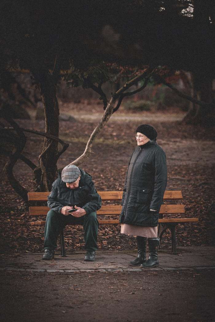 An elderly couple experiences a quiet moment in a park with the man seated on a wooden bench looking at his mobile phone, while the woman stands beside him looking away thoughtfully. A tangle of bare, dark branches creates a moody backdrop, suggesting the late autumn or winter season. The scene conveys a sense of stillness and the complexity of human relationships.