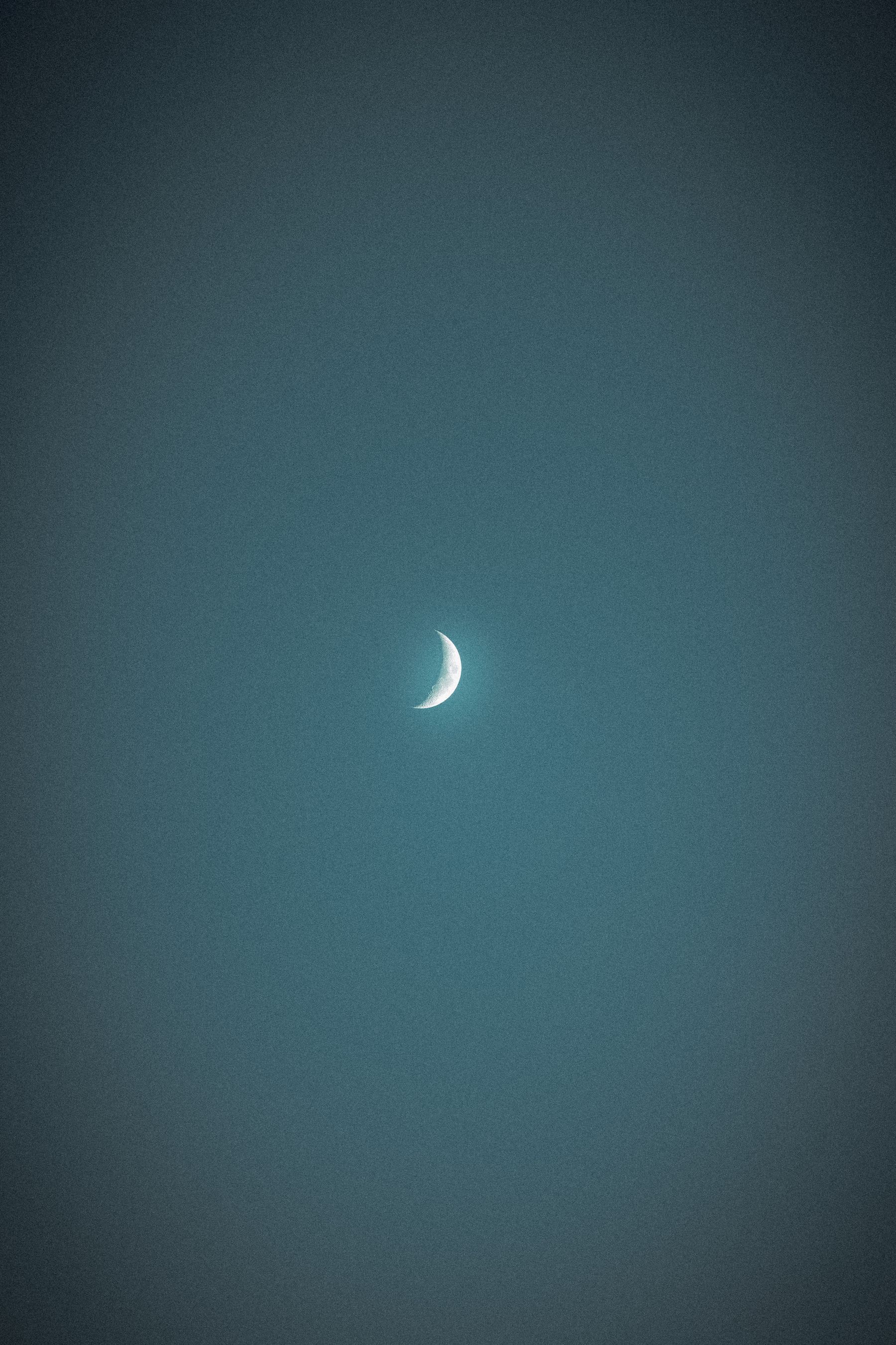 The image captures a waxing crescent moon centered in a dark, textured night sky. The moon is brightly lit on its curved edge, fading into darkness toward the rest of its surface, which is barely visible against the backdrop of the evening glow.