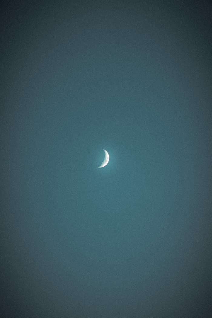 The image captures a waxing crescent moon centered in a dark, textured night sky. The moon is brightly lit on its curved edge, fading into darkness toward the rest of its surface, which is barely visible against the backdrop of the evening glow.