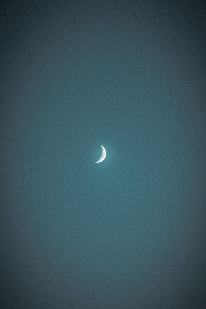 The image captures a waxing crescent moon centered in a dark, textured night sky. The moon is brightly lit on its curved edge, fading into darkness toward the rest of its surface, which is barely visible against the backdrop of the evening glow.