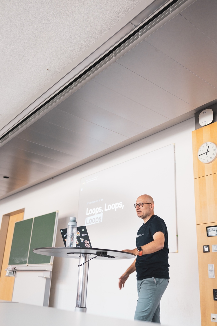 Florian standing at a small round table with a sticker-covered laptop, mid-sentence, in front of a projected slide reading 'Loops, Loops, Loops'.