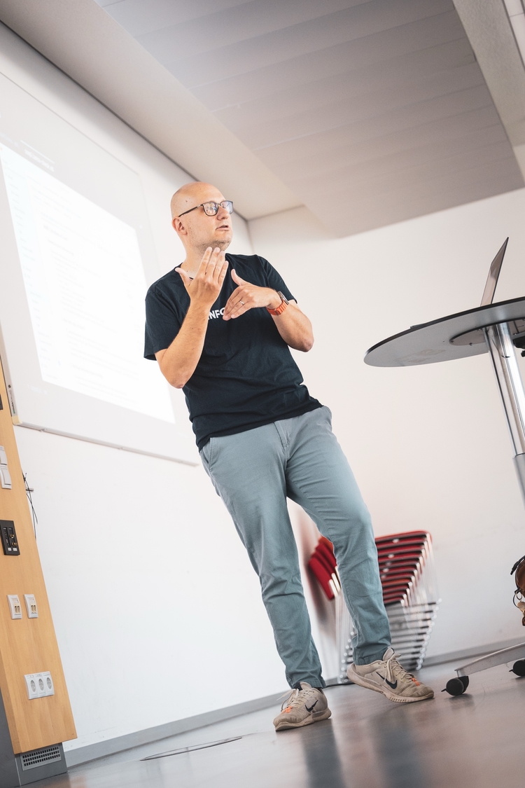 Florian gesturing with both hands while explaining a densely written slide projected on the wall behind him in a bright seminar room.
