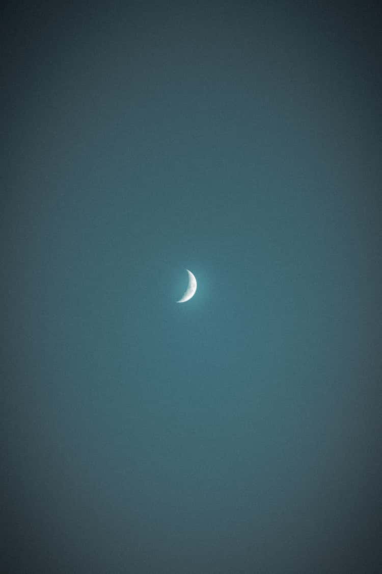 The image captures a waxing crescent moon centered in a dark, textured night sky. The moon is brightly lit on its curved edge, fading into darkness toward the rest of its surface, which is barely visible against the backdrop of the evening glow.