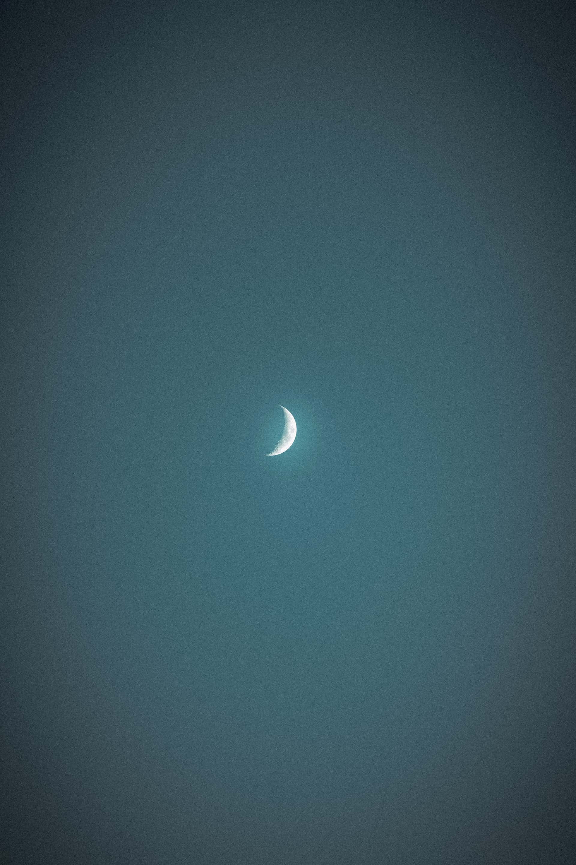 The image captures a waxing crescent moon centered in a dark, textured night sky. The moon is brightly lit on its curved edge, fading into darkness toward the rest of its surface, which is barely visible against the backdrop of the evening glow.