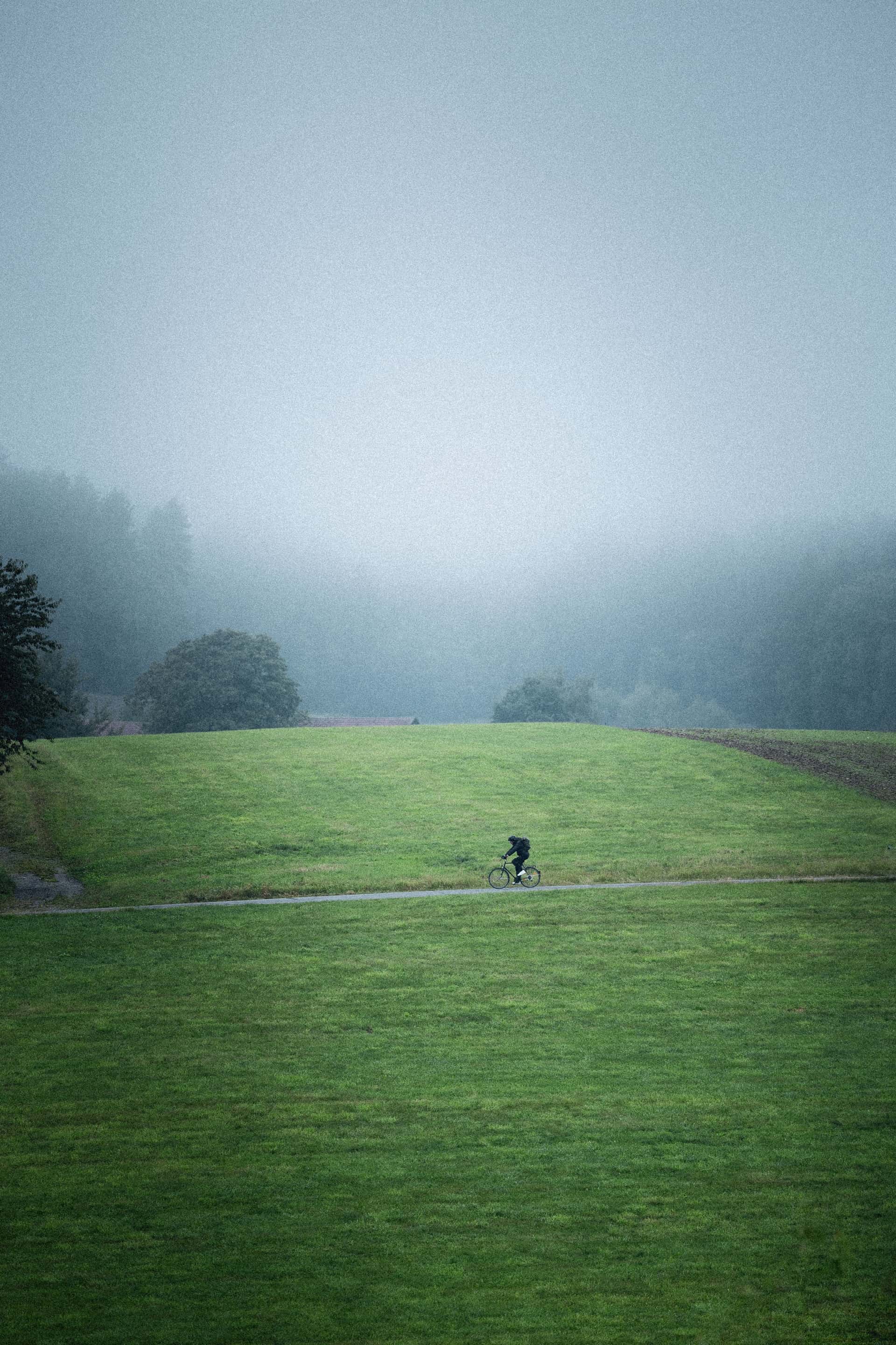 A lone cyclist rides along a narrow path through an expansive green field. The surroundings are enveloped in a thick mist, creating a serene and slightly mysterious atmosphere. In the background, the contours of trees and distant hills are barely visible through the fog, enhancing the sense of solitude and tranquility.