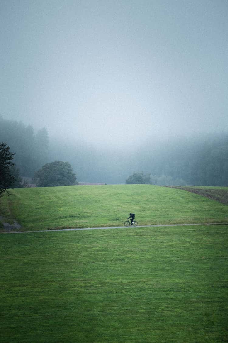 A lone cyclist rides along a narrow path through an expansive green field. The surroundings are enveloped in a thick mist, creating a serene and slightly mysterious atmosphere. In the background, the contours of trees and distant hills are barely visible through the fog, enhancing the sense of solitude and tranquility.