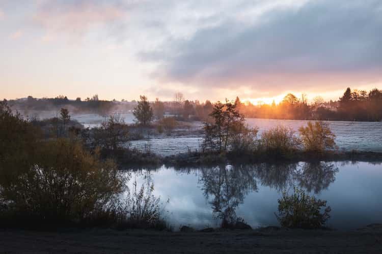 A tranquil landscape at dawn, featuring a still river reflecting the soft hues of sunrise. Frost covers the foreground and surrounding fields, while trees adorned with autumn leaves line the banks. In the background, gentle hills rise under a sky filled with soft clouds, bathed in golden light as the sun begins to rise. The overall atmosphere evokes a sense of peace and the beauty of nature's early hours.