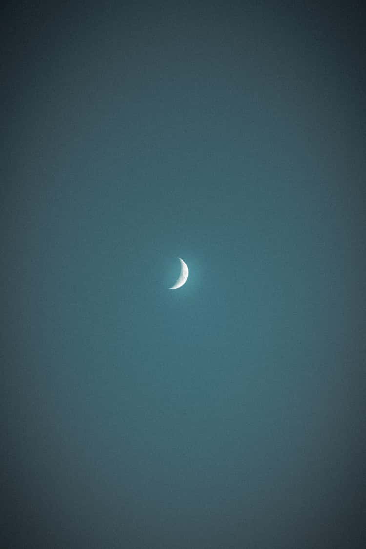 The image captures a waxing crescent moon centered in a dark, textured night sky. The moon is brightly lit on its curved edge, fading into darkness toward the rest of its surface, which is barely visible against the backdrop of the evening glow.