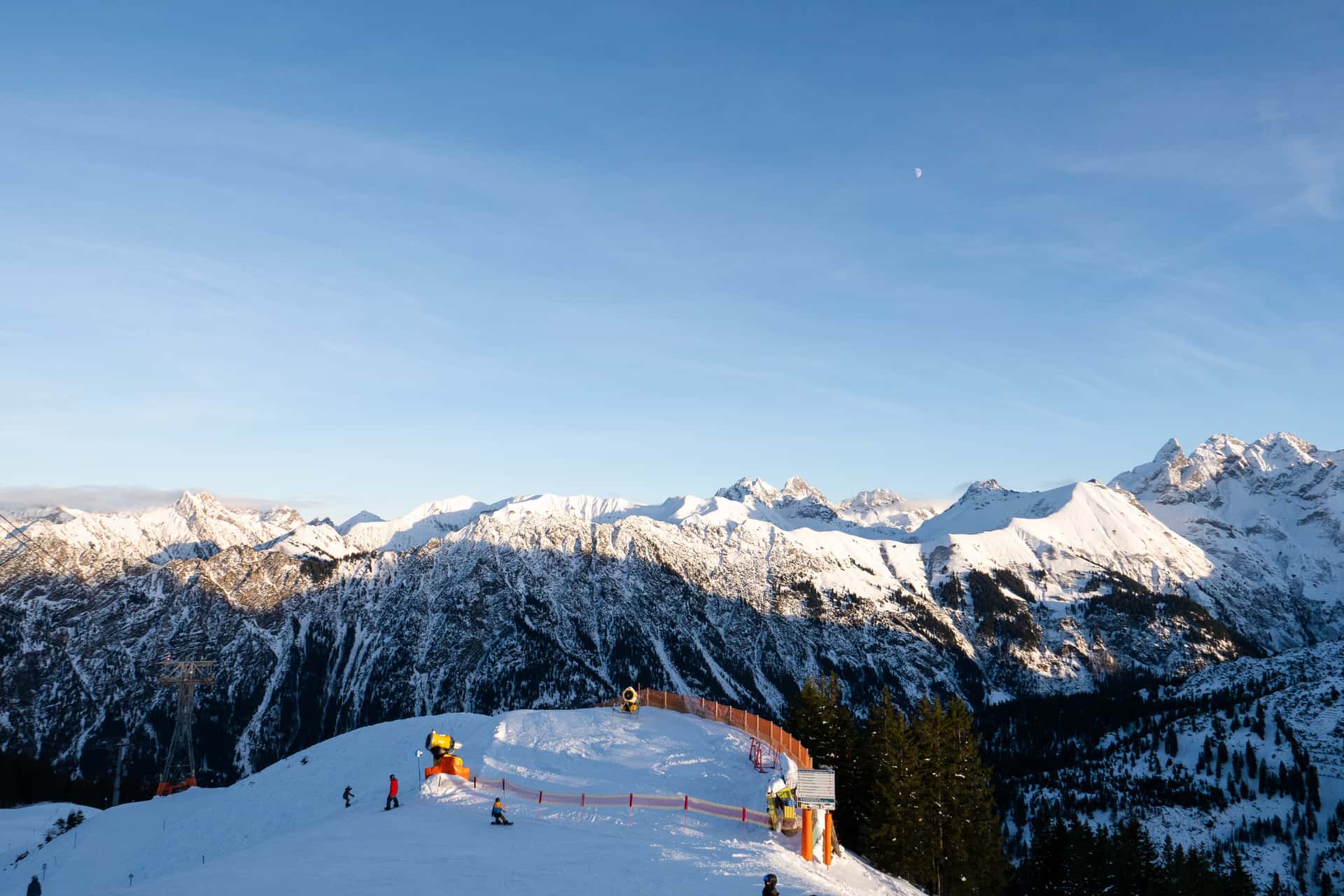 Snow-covered mountain slopes with skiers moving along a groomed trail near a curved orange safety fence, set against a backdrop of tall, rugged peaks coated in fresh snow under a clear sky with soft evening light.