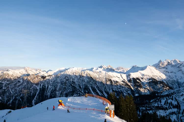 Snow-covered mountain slopes with skiers moving along a groomed trail near a curved orange safety fence, set against a backdrop of tall, rugged peaks coated in fresh snow under a clear sky with soft evening light.