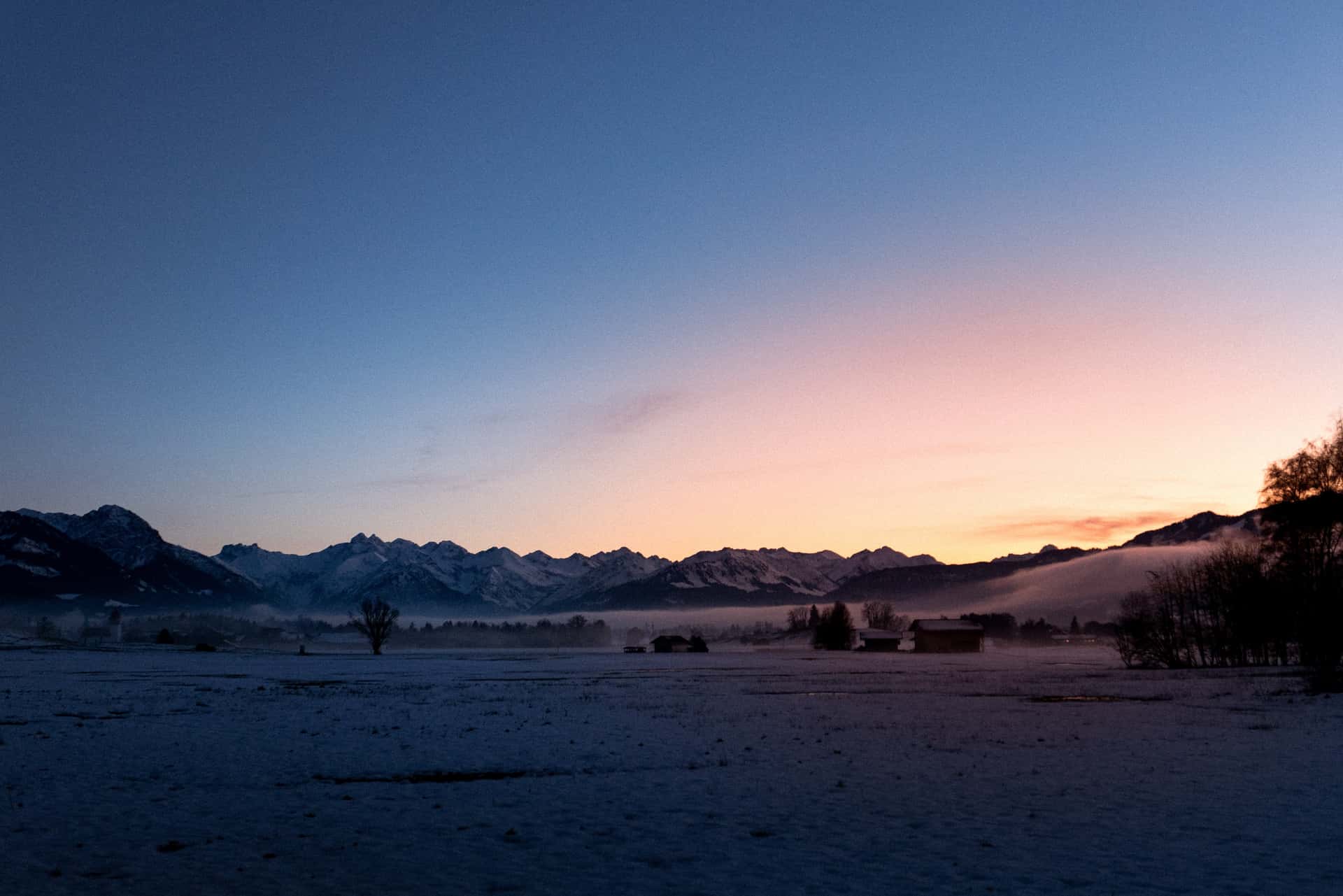 Snow-covered fields stretch toward a distant mountain range under a sky shifting from warm pink to deep blue as the sun sets. Soft mist lingers at the base of the mountains and around scattered trees and small buildings. The scene is calm and still, with the fading light of dusk illuminating the landscape.