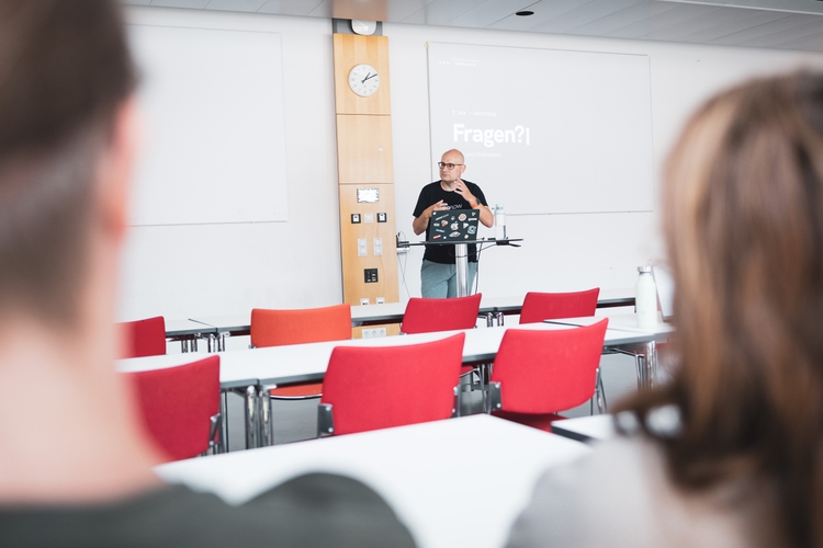 Florian speaking at a podium in a lecture hall at the University of Bayreuth, gesturing next to a laptop covered in stickers, with a terminal-styled slide reading "Fragen?" projected behind him and listeners in the foreground.