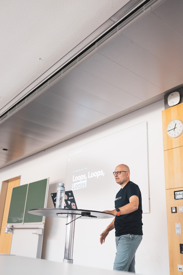 Florian standing at a small round table with a sticker-covered laptop, mid-sentence, in front of a projected slide reading 'Loops, Loops, Loops'.